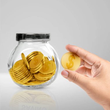 A human hand holding golden coin with golden coins in a glass jar on a white table. Financial concept