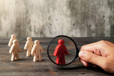 Magnifying glass focuses on a red wooden figure from the crowd of wooden figures on a wooden table. Leadership concept