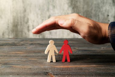 A human hand protects a red wooden figure from the two wooden figures on a wooden table. Leadership concept