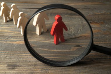 Magnifying glass focuses on a red wooden figure from the crowd of wooden figures on a wooden table. Leadership concept