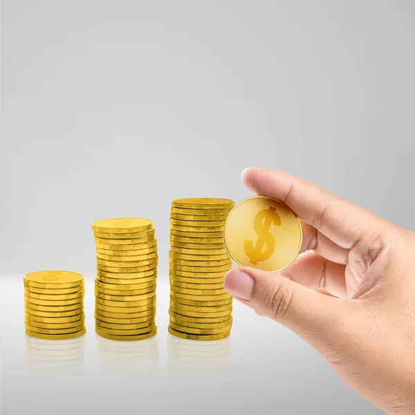 A human hand holding a golden coin with stairs of golden coins on a white table. Financial concept