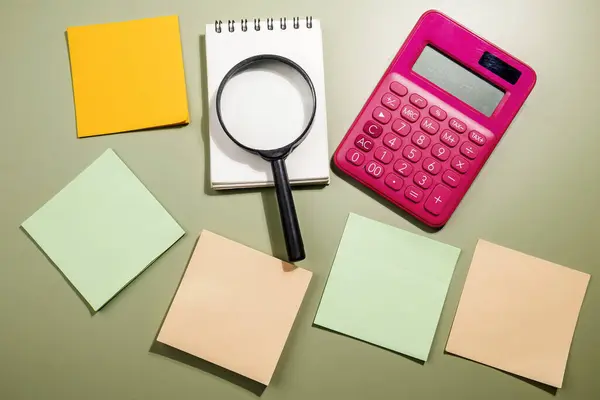 Closeup view of magnifying glass and calculator with an empty notebook and note paper on a colored background. Empty notebook and note paper for copy space