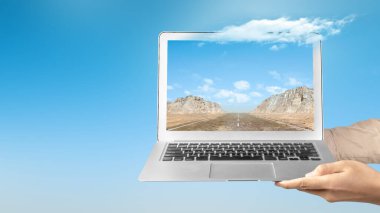 Tepeli bir cadde ve mavi gökyüzü olan bir kayalık manzaralı bir ekran manzaralı dizüstü bilgisayar tutan bir insan eli. Seyahat kavramı