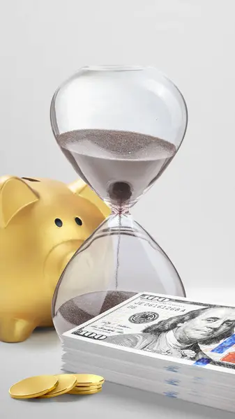 Close up hourglass and money agains golden piggy bank isolated over white background. Time is money concept.