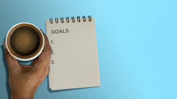 A notebook with goals list text and a cup of coffee on a colored background. Planning and target concept