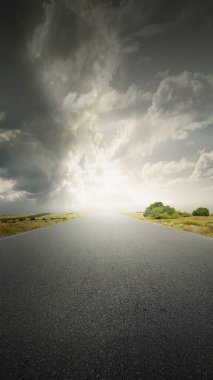 Asfalt yolu, yeşil çimlerle kaplı çayırlık alanda dramatik bir gökyüzü arka planı var.