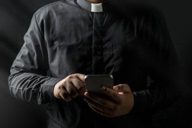 Genç rahip elinde akıllı telefon tutuyor ve siyah arkaplanda izole edilmiş ekrana dokunuyor..