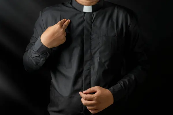 黒い背景に隔離された彼の体に十字架のサインをしている若い司祭 ロイヤリティフリーのストック画像