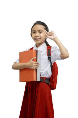 Sırt çantalı Asyalı bir ilkokul öğrencisinin portresi elinde kitap tutarken ve kafasını beyaz bir arka planda izole bir şekilde gösterirken.
