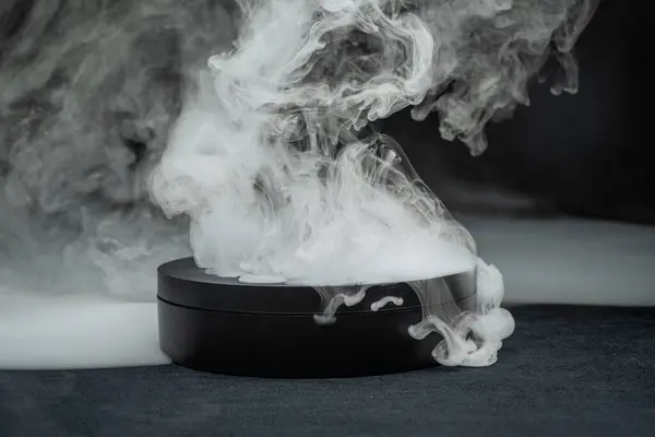 stock image White smoke is emanating from a round black box on a dark background. The smoke is billowing up and over the sides of the box