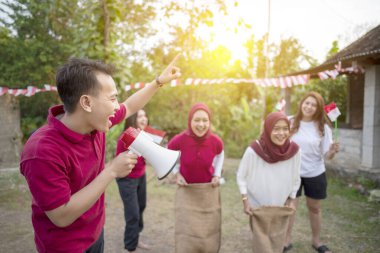 Kırmızı gömlekli bir adam bir megafon tutuyor ve gökyüzünü işaret ediyor. Etrafında bir grup insan dikiliyor, bazıları bayrak tutuyor. Sahne kutlama ve neşe doludur. Endonezya 'nın bağımsızlık günü kavramı
