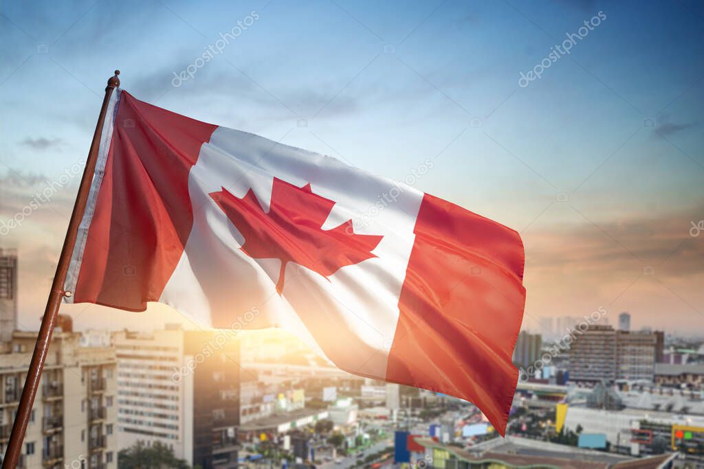 A red and white Canadian flag is flying high in the sky over a city. The flag is waving in the wind, and the sun is shining brightly on it, creating a warm and inviting atmosphere