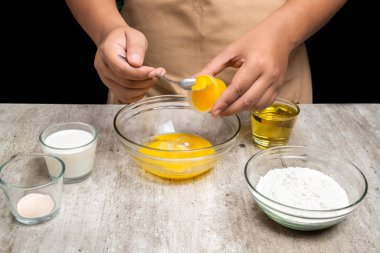 Bir insan masanın üzerinde bir kaseye yumurta kırıyor. Masanın üzerinde başka kase ve fincanlar var ve yakınlarda bir kaşık var.