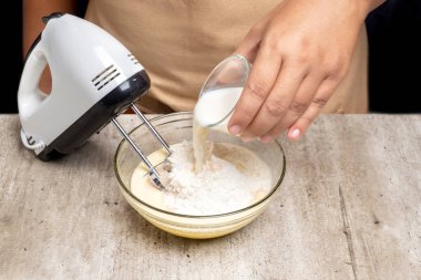 Bir insan mikser kullanırken bir kaseye süt döküyor. Kase un ve şekerle dolu.