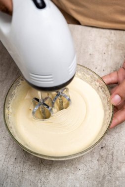 Bir kişi pasta yapıyor ve masanın üzerinde bir kase un ve şeker var. Malzemeleri karıştırmak için karıştırıcı kullanıyor.