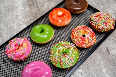 Bir tepsi rengarenk mini donut ve üzerine de şeker serpiştirilmiş. Mini çörekler arka arkaya diziliyor, bazıları yeşil, bazıları pembe. Tepsi tezgahın üstüne konmuş ve çörekler çok lezzetli görünüyor.