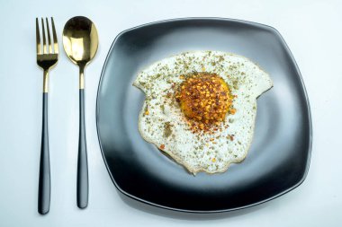 Bir tabak güneşli yumurta yemeği, siyah tabak, altın çatal ve kaşık. Tabakta biraz baharatlı sahanda yumurta var.