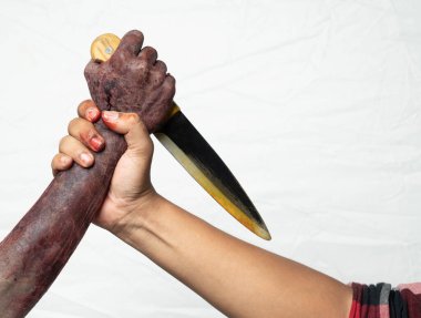 Beyaz bir arka plan için izole edilmiş bir adamla kanlı bir yarayla savaşan zombi bir el. Bıçak tutan zombi bir el ve zombi eli tutan bir adam. Bıçak insan eline doğrultulmuş, gergin ve tehlikeli bir atmosfer yaratıyor. Ürkütücü Cadılar Bayramı