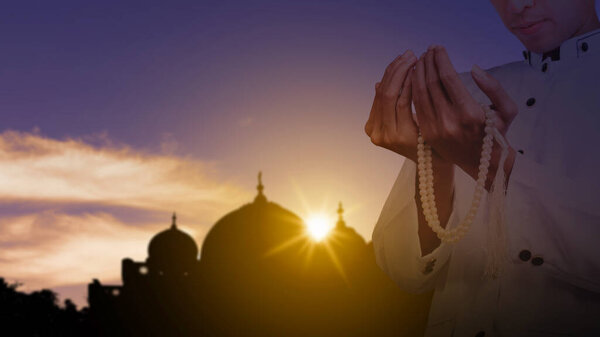 Muslim man praying with prayer beads with a view of the silhouette of the mosque with sunlight in the sunset scene background. Concept of Religion, Spiritual or Ramadan