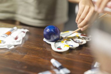 Küçük bir kız masanın üstünde paskalya yumurtası süslemesi için fırçayla yumurta boyuyor. Paskalya bayramını kutlama kavramı