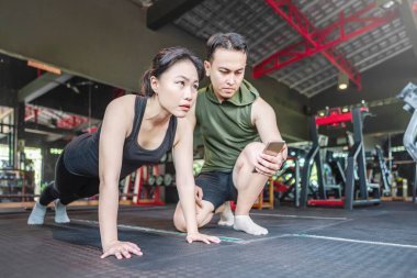 Endonezyalı Güneydoğu Asyalı özel antrenör spor salonundaki Çinli Doğu Asyalı kadına cep telefonuyla egzersiz adımları gösteriyor. Spor salonu ve sağlıklı yaşam tarzı konsepti