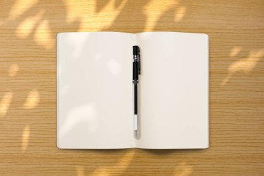 Açık bir defter ve tahta arka planda gölgesi olan bir kalem. Boş kağıt ya da yaprak. Tasarımınız için boşluk oluştur veya kopyala