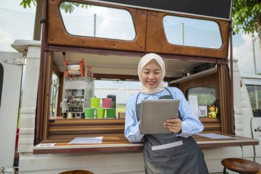 Endonezyalı güneydoğu Asyalı tesettürlü, tesettürlü Müslüman kadın barista kafesinin önünde tablet kullanırken ayakta duruyordu. Kamyon üzerinde küçük bir kafe ya da kafe işletmesi