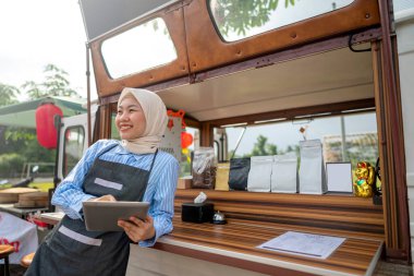 Endonezyalı güneydoğu Asyalı tesettürlü, tesettürlü Müslüman kadın barista kafesinin önünde tablet kullanırken ayakta duruyordu. Kamyon üzerinde küçük bir kafe ya da kafe işletmesi