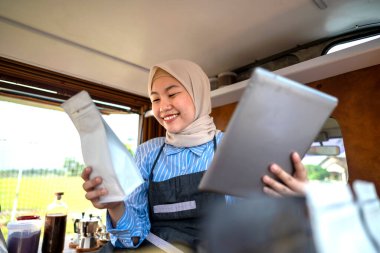 Endonezyalı güneydoğu Asyalı tesettürlü, tesettürlü Müslüman kadın barista kafesinde tablet kullanırken oturuyor. Kamyon üzerinde küçük bir kafe ya da kafe işletmesi