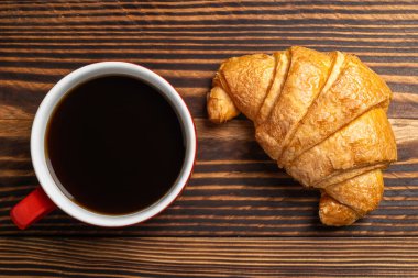 Ahşap zemin üzerinde bir fincan kahveyle taze pişmiş kruvasan. Üst görünüm