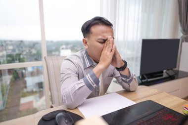 Endonezyalı güneydoğu Asyalı işadamı hibrit iş yerinde dizüstü bilgisayar üzerinde çalışırken stres ve tükenme yaşıyor. Evrak işleri ve masanın üstünde bir cep telefonu. İş konsepti. Evden uzak çalışma