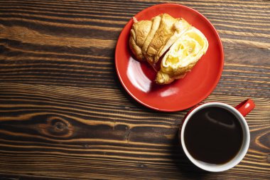 Kırmızı tabakta taze pişmiş kruvasan dilimi ve ahşap arka planda bir fincan kahve. Üst görünüm