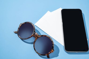 Beyaz kağıt, güneş gözlüğü ve mavi arka planda bir cep telefonu. Model veya Kopyala uzayı. Seyahat macerası, seyrüsefer ve planlama. Seyahat, turizm veya tatil kavramı