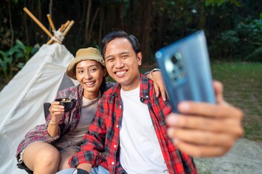 Endonezyalı güneydoğu Asyalı çift cep telefonuyla selfie çekiyor. Kadın telefonu tutar, yanında bir adam ve bir kase taşır. Doğal ortamdaki yemekten zevk alma sahnesi.