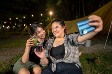 Endonezyalı Güneydoğu Asyalı kadınlar cep telefonlarını açık havada birlikte kullanarak selfie çekiyorlar ve içeceklerin tadını çıkarıyorlar. Doğal ortamdaki yemekten zevk alma sahnesi.