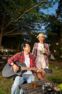 Endonezyalı güneydoğu Asyalı bir adam arkadaşlarıyla dışarıda akustik gitar çalıyor. Rahat ve keyifli bir yaşam tarzı gösteriyorum. Kamp yapmanın doğal ortamında keyif alma sahnesi.