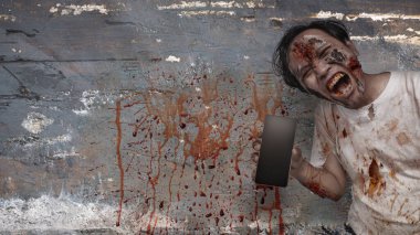 Endonezyalı güneydoğu Asyalı yaralı ve duvarında kan lekeleri olan bir cep telefonu tutan korkunç erkek zombi. Model veya Kopyala uzayı. Sahne korkutucu ve ürkütücü. Ürkütücü Cadılar Bayramı kavramı