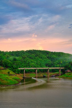 Huzurlu bir nehri kaplayan, nefes kesici bir günbatımı ile yemyeşil tepelerin üzerine sıcak bir parıltı saçan bir köprünün resmedilmiş görüntüsü. Görüntü bir huzur ve sükunet hissi uyandırıyor..