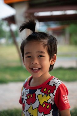 Kendine özgü saç stili olan genç bir çocuk parkta oynarken parlak bir şekilde gülümsüyor. Yeşillik ve açık hava oyun alanı neşeli bir atmosfer yaratır..