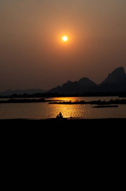 Güneş, sulara turuncu ve altın renkler dökerek huzurlu bir gölün üzerinde yavaşça batarken, karanlık dağ siluetleri manzaraya çarpıcı bir zemin oluşturur..