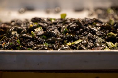 Buharda pişirilmiş siyah midyeler, taze otlarla süslenmiş bir satıcının tepsisinde üst üste oturur, öğleden sonra güneş ışığında deniz ürünleri pazarında canlı bir seçim sergilerler, arka planda müşteriler vardır..