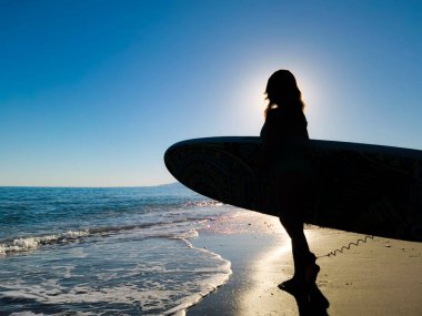 A captivating silhouette of a woman holding a surfboard, captured against the setting sun on the beach. The surfboard's intricate designs are subtly visible, while the calm ocean and clear sky create a serene and dreamy backdrop. The woman's outline  clipart