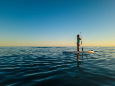 Yeşil mayo giymiş bir kadın, gün batımında açık denizlerin sakin sularında usulca kürek çekiyor. Gökyüzü, dalgalanık su yüzeyini yansıtan, altından yumuşak maviye dönüşür. Huzurlu sahne, barış yandaşlarını yakalar.