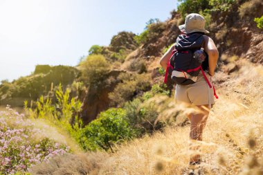 Yeşil ve çiçeklerle çevrili güneşli dağ arazisinde yürüyüş yapan şapkalı ve sırt çantalı bir kadın. Açık hava maceralarını ve doğa keşiflerini yakalar..