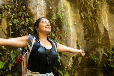 A woman stands with arms outstretched under a refreshing waterfall in a lush canyon. She is wearing a backpack and appears to be on a hiking adventure, soaking in the natural beauty. clipart