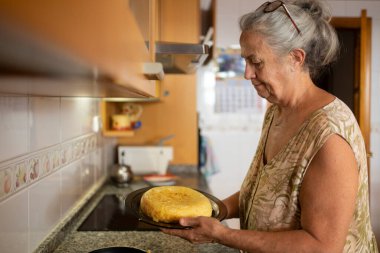 Mutfakta yeni pişmiş İspanyol tortillasını tutan desenli elbiseli yaşlı bir kadın..