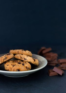 Çikolata parçalı kurabiyeler ve bir tabakta çikolata parçaları ve koyu bir arka plan.