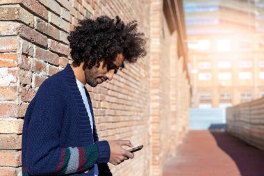 Portrait of young afro man using his mobile phone against brick wall. Technology and lifestyle concept.