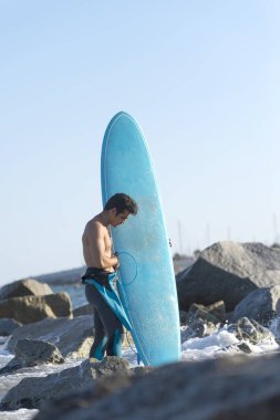 Genç sörfçünün çıplak ayakla kumlu sahilde durduğu engin okyanusa bakan ve sörf tahtasını tutan rüzgarlı yaz gününde dalgalara çarpmaya hazır sörfçünün arkası.