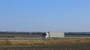 Güneşli bir havada beyaz bir araba otoban boyunca sürer. Otoyolda kamyon var..